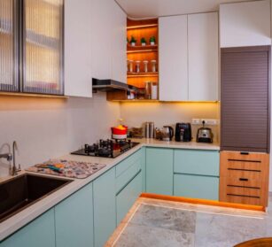 Modern L-shaped modular kitchen with wood finish cabinets, built-in appliances, glass overhead units, and natural lighting