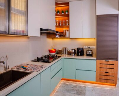 Modern L-shaped modular kitchen with wood finish cabinets, built-in appliances, glass overhead units, and natural lighting